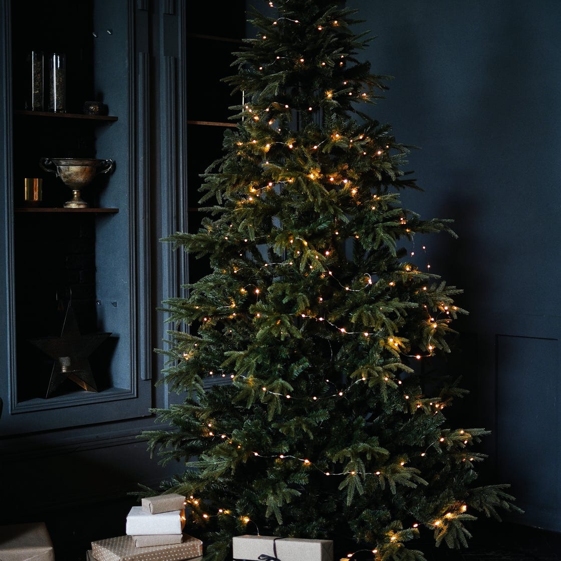 Tall green Christmas tree decorated with small glowing garlands and surrounded by shelves and bunch of boxes with gifts in dark room